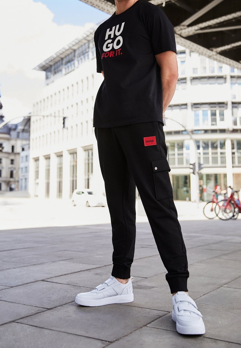 Homme portant un t-shirt noir "HUGO FOR IT.", un pantalon cargo noir avec un patch rouge Hugo et des baskets blanches, debout sur un trottoir en ville.