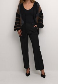 Cardigan oversize a maglia con strisce nere, marroni e grigie; abbinato a pantaloni neri a vita alta e décolleté nere a punta.