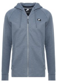 Šedá mikina na zip vyrobená z jemné tkaniny, s předním zipem, dvěma bočními kapsami a malou černou nášivkou na horní levé straně.