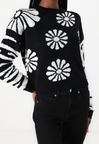 Black knitted sweater featuring white floral and striped patterns, ribbed collar and hem. Worn with black jeans, showcasing a relaxed fit.