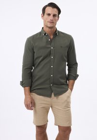 Chemise boutonnée olive à manches longues, arborant un logo discret et des boutons blancs, associée à un short beige en tissu léger.