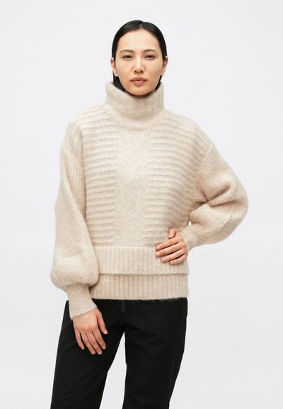 Kvinde står iført en beige, ribbet, højhalset sweater med ballonærmer og sorte bukser foran en ensfarvet lys baggrund.