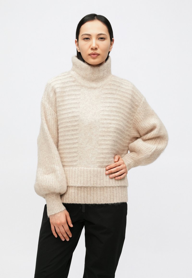 Donna in piedi che indossa un maglione beige a coste con collo alto e maniche a palloncino, abbinato a pantaloni neri, davanti a uno sfondo chiaro e uniforme.