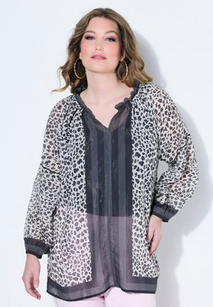 Nainen pukeutuneena läpikuultavaan mustavalkoiseen leopardikuosiseen puseroon, jossa röyhelökaulus ja suuret rengaskorvakorut, poseeraa valkoista taustaa vasten.