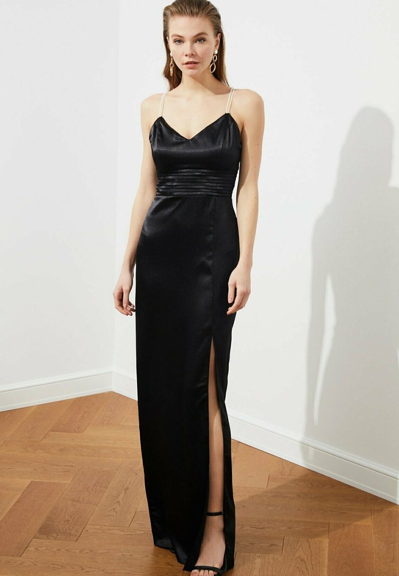 Kobieta w czarnej satynowej sukni wieczorowej z perłowymi ramiączkami i rozcięciem bocznym, stojąca na drewnianej podłodze przy białej ścianie.