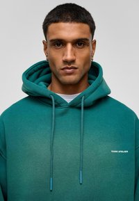 Teal hoodie met een geleidelijke kleurverloop, voorzien van een frontzak, trekkoorden met blauwe uiteinden en een wit "YOUNG ATELIER" logo.