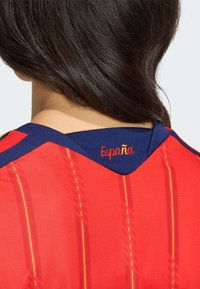 Punane spordisärk v-kaelusega ja tumesiniste detailidega, kaetud kollaste vertikaalsete triipudega ning selja kaelusel tikitud sõnaga "España".