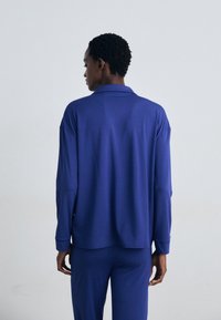 Chemise à manches longues en tissu bleu uni avec un col, coupe décontractée et ourlet arrière légèrement plus long, assortie à un pantalon bleu.