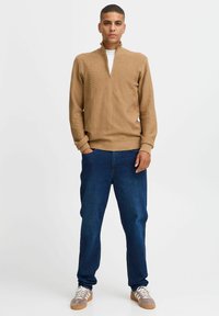 Khaki gestrickter Pullover mit hohem Kragen, kombiniert mit dunkelblauen Jeans. Strukturierter Stoff, lockere Passform und lässige Schnürschuhe mit Gummisohle.