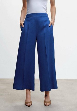 Mujer con pantalones azules de talle alto y pierna ancha con pliegues delanteros, camiseta blanca sin mangas y tacones abiertos marrones, de pie con las manos en los bolsillos.