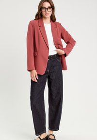 Rosenfarbener Blazer mit taillierter Passform, getragen über einem weißen Top, kombiniert mit dunkelblauen hochgeschnittenen Jeans und schwarzen Sandalen.