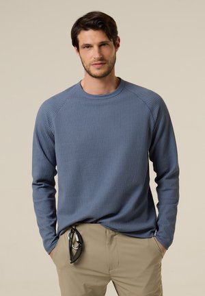 Uomo con capelli scuri che indossa una camicia a maniche lunghe blu a trama e pantaloni beige, con occhiali da sole appuntati alla tasca, in piedi con le mani in tasca.