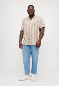 Camisa de manga corta y botones en color marrón claro con rayas verticales blancas, combinada con jeans azul claro y zapatillas blancas. Cuello plano y corte relajado.