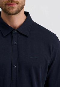 Een bebaarde man die een marineblauwe knopenhemd met kraag en het "Pierre Cardin" logo op de borst draagt, tegen een eenvoudige achtergrond.