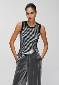 Haut sans manches à rayures noires et blanches avec des boutons dorés sur les épaules, associé à un pantalon large gris brillant avec poches avant.