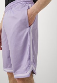 Lilla træningsshorts med et struktureret stof, elastisk talje, sidelommer og hvide accentstriber langs siderne.