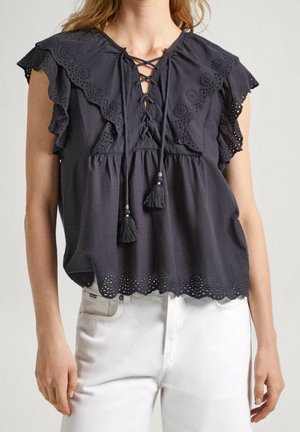 Černá bezrukávová halenka s šněrováním vpředu, krajkovými volánky na ramenou a vroubkovaným lemem, nošená s bílými kalhotami.