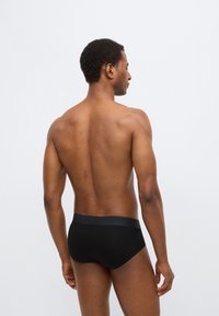 Zwarte katoenen slips met een brede elastische tailleband. De stof is glad en sluit nauw aan. Het model wordt van achteren getoond.