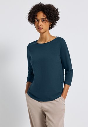Haut à manches longues bleu canard avec un col rond, en tissu léger et détails de couture sur les côtés. Associé à un pantalon de couleur claire.