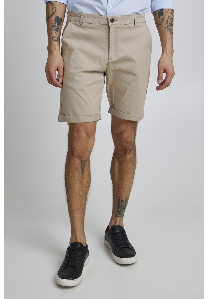 Pantalones cortos de algodón beige con los dobladillos enrollados, con cierre de botón y bolsillos, usados con zapatillas negras. Tatuajes visibles en las piernas.
