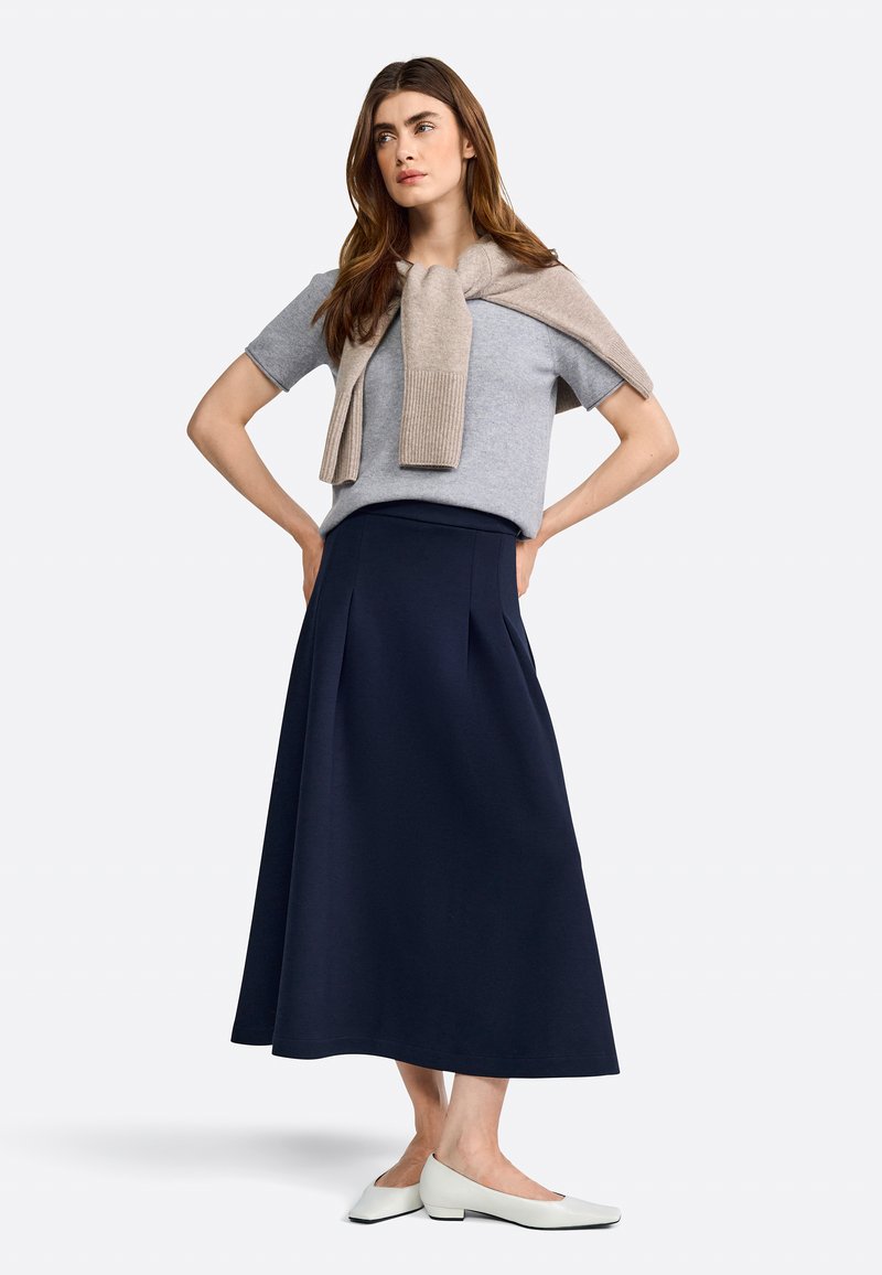 Suéter gris de manga corta con una bufanda beige caída sobre los hombros, combinado con una falda larga de color marino y zapatos blancos de tacón bajo.