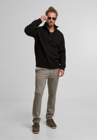 Zwarte ritsoverzameling, beige broeken en grijze suède schoenen. Bril accentueert de look. Stijl is casual en samenhangend in kleur.