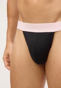 Czarny bawełniany thong z szerokim, jasnoróżowym paskiem na biodra, na którym subtelnie umieszczono logo "Calvin Klein". Gładka tekstura i dopasowany krój.