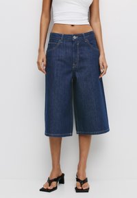 Culottes en denim bleu foncé avec une taille haute. Dotées de poches avant et de coutures visibles. Texture lisse. Portées avec des sandales à talons noires.