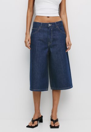 Culottes de mezclilla azul oscuro con cintura alta. Presentan bolsillos frontales y costuras visibles. Textura suave. Combinados con sandalias negras de tacón.