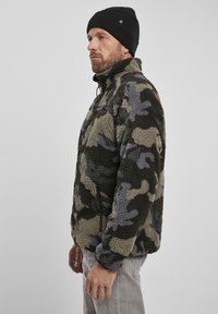 Fleece jacket in camouflage pattern with shades of green, gray, and black. Features a high collar and zippered pocket. Soft texture.