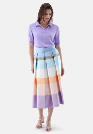 Donna che indossa una polo a maniche corte color lavanda, una gonna plissettata a vita alta con righe orizzontali pastello e sandali bianchi con tacco.