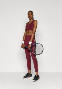 Sporta apģērbs, kas sastāv no tumši sarkanas sporta krūštura un saskaņotiem legingiem ar ziedu akcentu sānos, kopā ar melnā krāsā tenisēm.