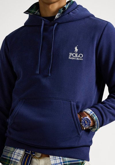 Μπλε σκούρο φούτερ με τσέπη μπροστά, κουκούλα με κορδόνι και κεντημένο λογότυπο "Polo Ralph Lauren". Συνδυάζεται με γιακά πουκάμισου καρό από κάτω.