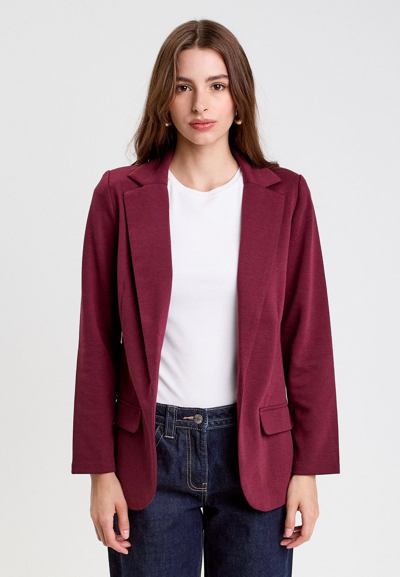 Burgundy blazer with notched lapels, two front pockets, and long sleeves. Worn over a white shirt, shown with dark blue jeans. Smooth texture.
