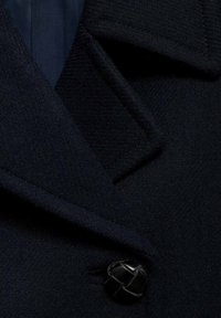 Tummansininen teksturoitu takkuteksti, jossa on lähikuva mustasta napista ja terävistä kauluksista, korostaen kudottujen herringbone-kuvioiden yksityiskohtia.