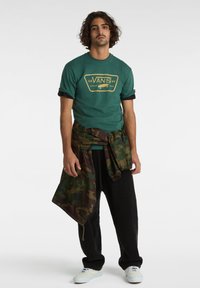 Camiseta verde con logo amarillo de Vans, combinada con pantalones de pana negros. Chaqueta de camuflaje atada a la cintura, zapatillas blancas. Luz natural, fondo simple.