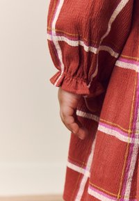 Vestito a quadri rosso con strisce bianche, rosa e gialle. Caratterizzato da un tessuto strutturato e manica arricciata con orlo a volant. Mano che tiene il vestito.