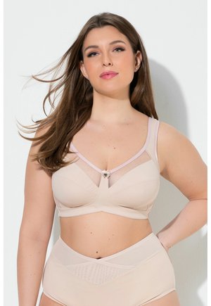 Kvinde med langt brunt hår iført bh i nøgenfarvet og højtaljede trusser, stående mod en hvid baggrund med hånden på hoften.