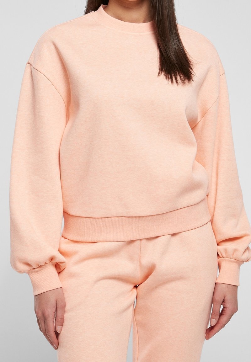 Sweat-shirt rose clair à encolure ronde, épaules tombantes et manches froncées, fabriqué en tissu doux. Assorti à un pantalon de jogging rose assorti.