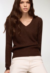 Maglione marrone in maglia con scollo a V, maniche lunghe e un motivo testurizzato, abbinato a pantaloni chiari.