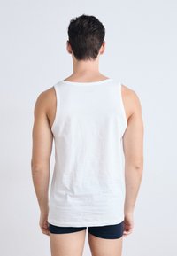 Balta kokvilnas bezpiedurkņu krekls ar apaļu kakla izgriezumu, izceļošs gludu tekstūru un brīvu piegriezumu. Valkāts kopā ar tumši ziliem šortiem, skatoties no aizmugures.