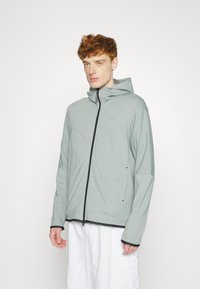 Jong iemand met krullend rood haar, gekleed in een lichtgrijze Nike zip-up hoodie en losse witte broek, staande tegen een plain witte achtergrond.