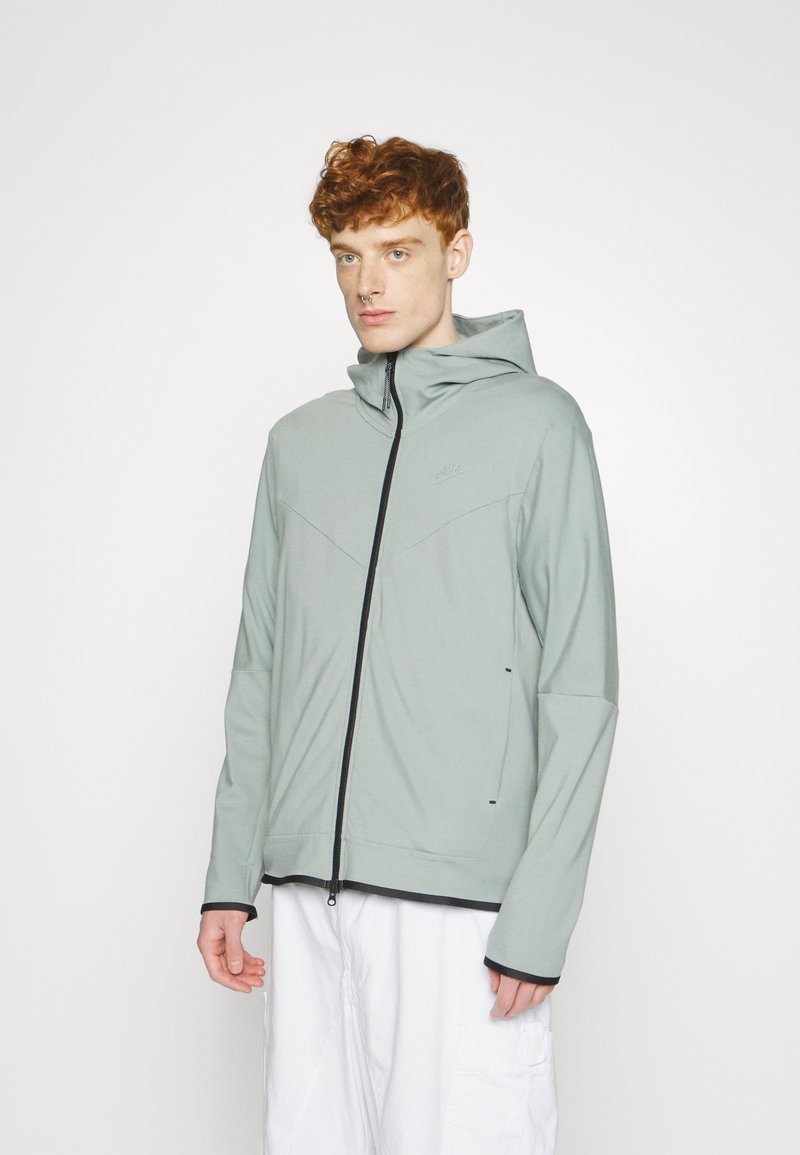 Jeune personne avec des cheveux roux bouclés, portant un sweat à capuche gris clair zippé de Nike et un pantalon blanc ample, se tenant contre un fond blanc uni.