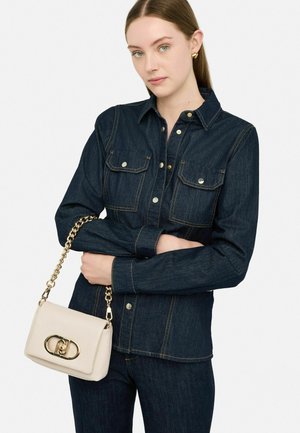 Camicia button-up in denim scuro con due tasche sul petto, accentuata da cuciture dorate. Una borsa beige chiara con tracolla in catena dorata.