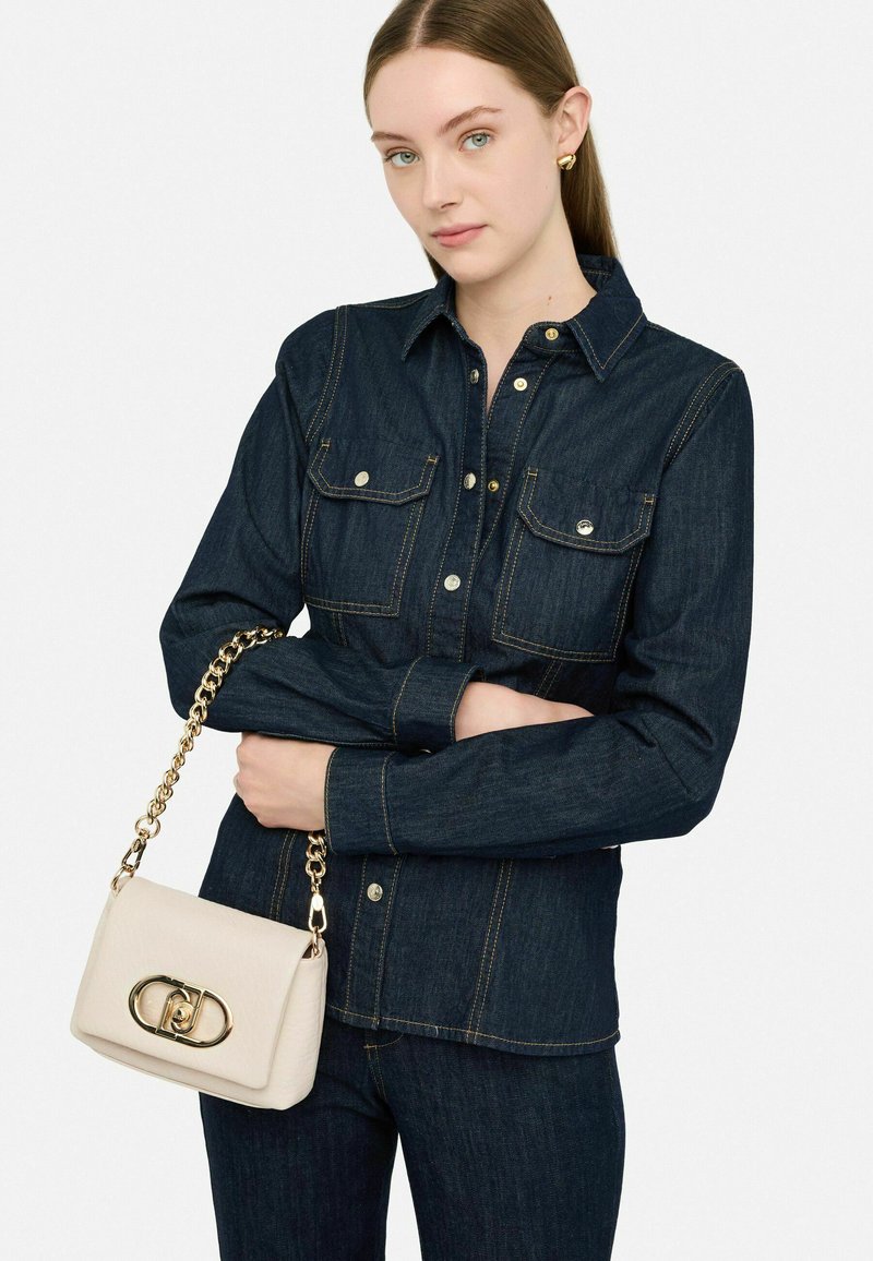 Camicia button-up in denim scuro con due tasche sul petto, accentuata da cuciture dorate. Una borsa beige chiara con tracolla in catena dorata.