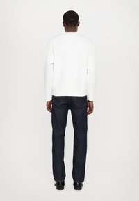 Biała bluza z okrągłym dekoltem, ściągaczami przy rękawach i u dołu, w połączeniu z ciemnoniebieskimi jeansami. Model widziany od tyłu.