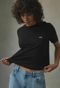 Černé bavlněné tričko s krátkým rukávem, které má nad lemem malé bílé vyšívané text "self.", v kombinaci se světle modrými denimovými džíny.
