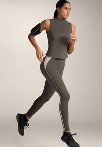 Atletische vrouw die jogt in een taupekleurige mouwloze top met rits, bijpassende legging met witte zijpanelen, zwarte sneakers en een armband voor haar telefoon.
