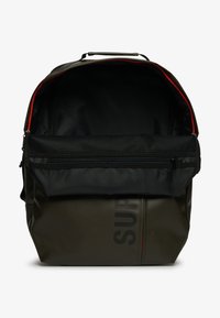 Ciemnozielony plecak z górnym uchwytem, otwarta główna komora pokazująca czarne wnętrze i czerwone wykończenie zamka, oraz przednia kieszeń z siatki zapinana na zamek.