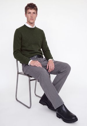 Homme assis sur une chaise en métal portant un pull vert, une chemise blanche, un pantalon gris, une ceinture noire et des bottes noires sur fond blanc.